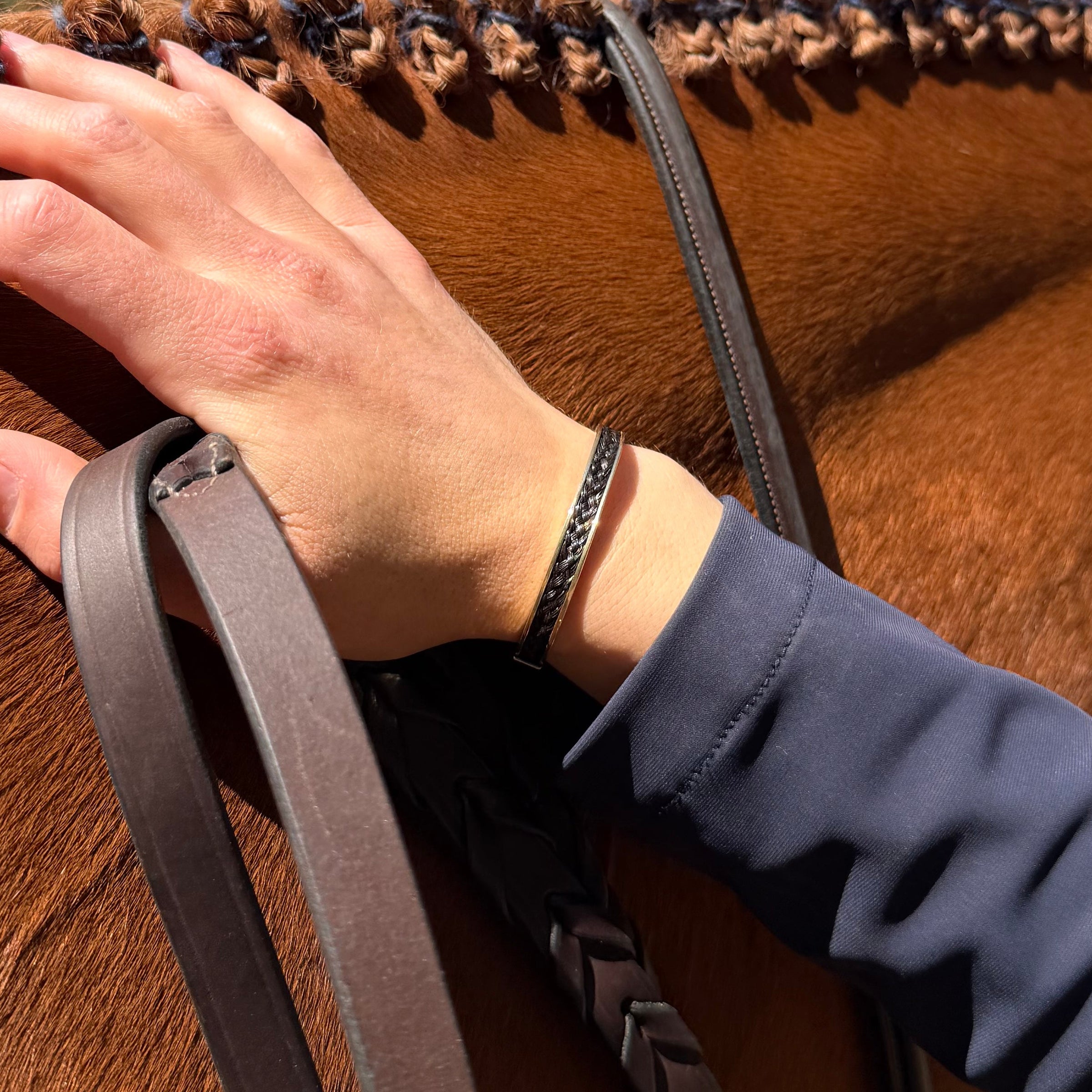 Equestrian wearing a gold horsehair cuff bracelet while holding reins against a chestnut horses neck with show braids.